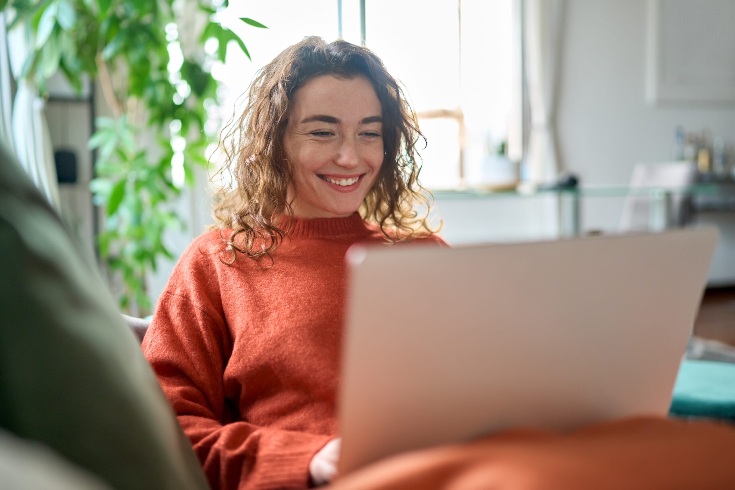 Eine junge Frau in einem roten Pullover sitzt lächelnd da und hat ihren Laptop auf dem Schoss.